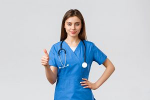 CNA wearing blue scrubs and stethoscope while smiling and giving thumbs up sign.