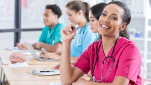 a female nurse smiles at the camera with other medical students in training out of focus in a classroom with nurses and med techs