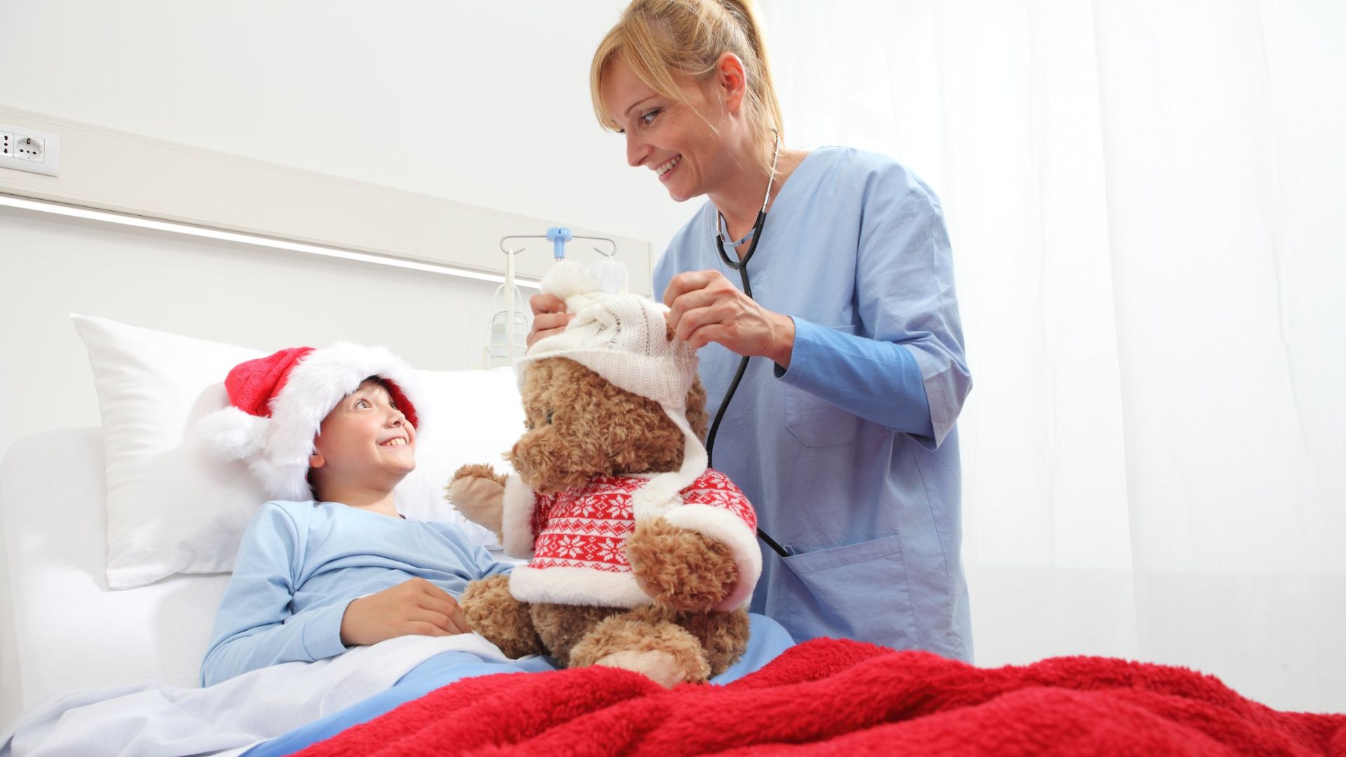 a nurse smiles taking care of a smiling child patient in the hospital room by bringing in a holiday themed teddy bear