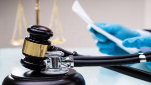 a black gavel and stethoscope sit on a table with a out of focus person wearing blue nitrile jobs looking over malpractice insurance paperwork