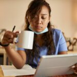 A tired nurse pores over her notes on her tablet, holding a cup of coffee in her hand. Image demonstrates the long hours required in some nursing jobs.