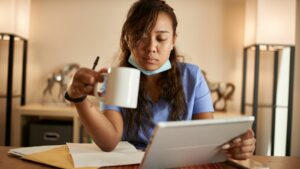 A tired nurse pores over her notes on her tablet, holding a cup of coffee in her hand. Image demonstrates the long hours required in some nursing jobs.