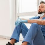 A male nurse leans back against a white window and wall, arms crossed over his legs, mask off. He looks tired. Image demonstrates how demanding RN jobs can be.