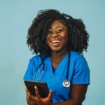 A nurse practitioner stands against a plain background, smiling and holding a device.
