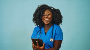 A nurse practitioner stands against a plain background, smiling and holding a device.