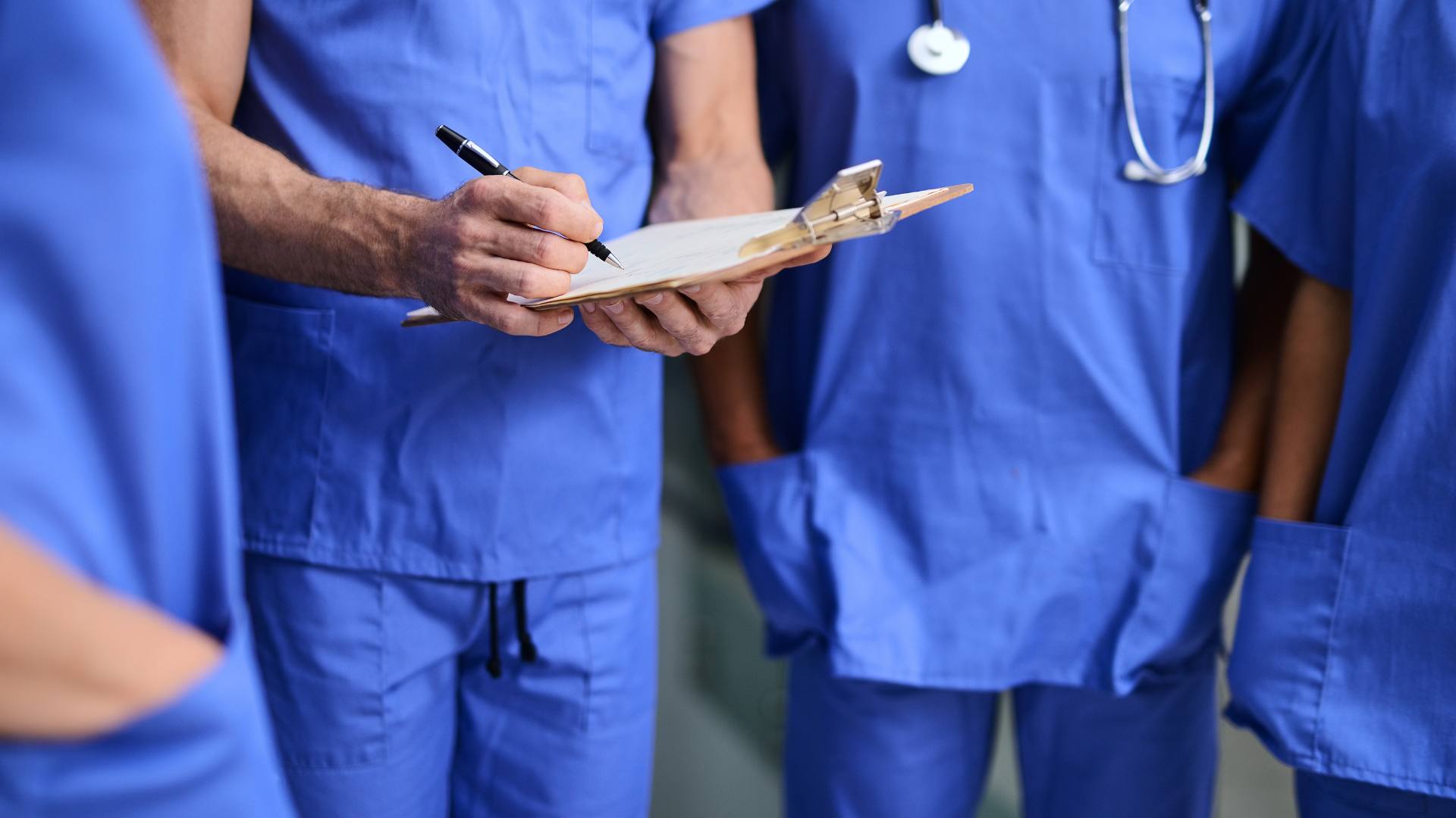 Four nurses wearing blue scrubs stand together. One writes on a clipboard. This is demonstrating the transition from travel nursing.
