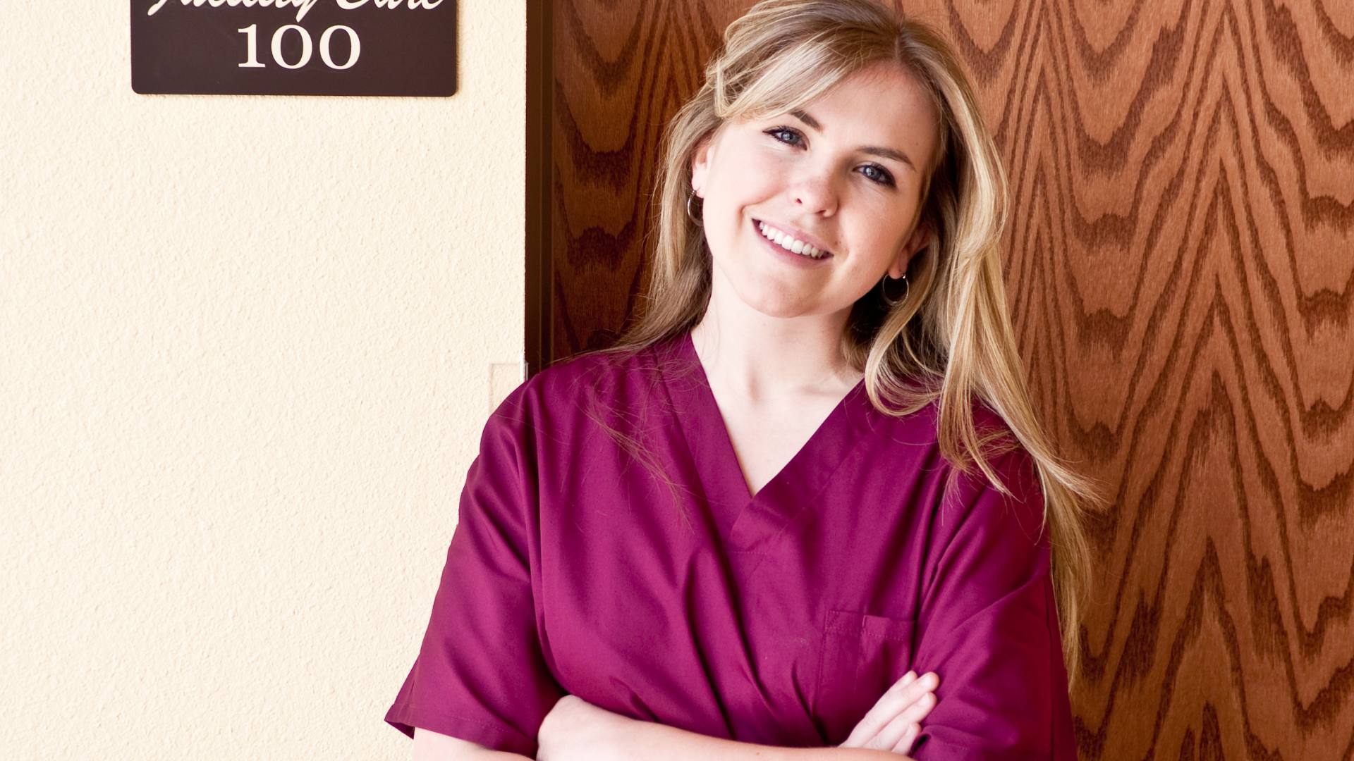 A blonde woman stands with her arms crossed and smiles at the camera while wearing maroon scrubs, demonstrating the difference between an lpn vs rn.