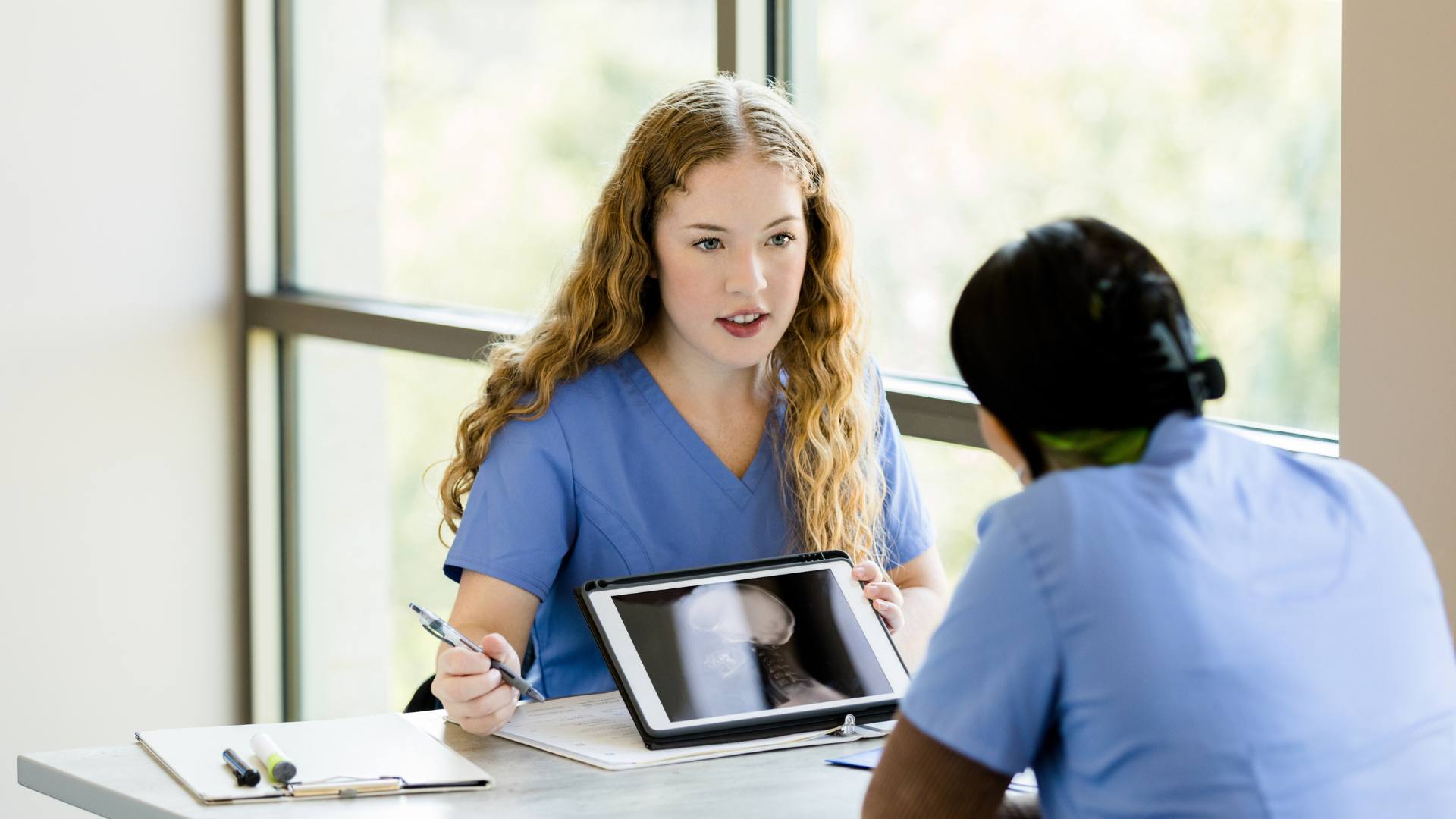 A med tech wearing blue scrubs shows an x-ray scan on an iPad to a nurse, sitting across from her.