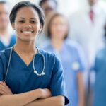 A nurse wears blue scrubs and a stethoscope around her neck while smiling with her arms crossed, demonstrating successful nursing placement.