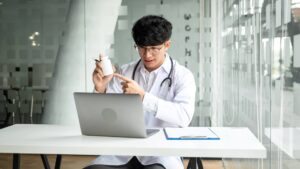 A person wearing a white lab coat sits at a desk and holds up a medication bottle, demonstrating med tech classes.